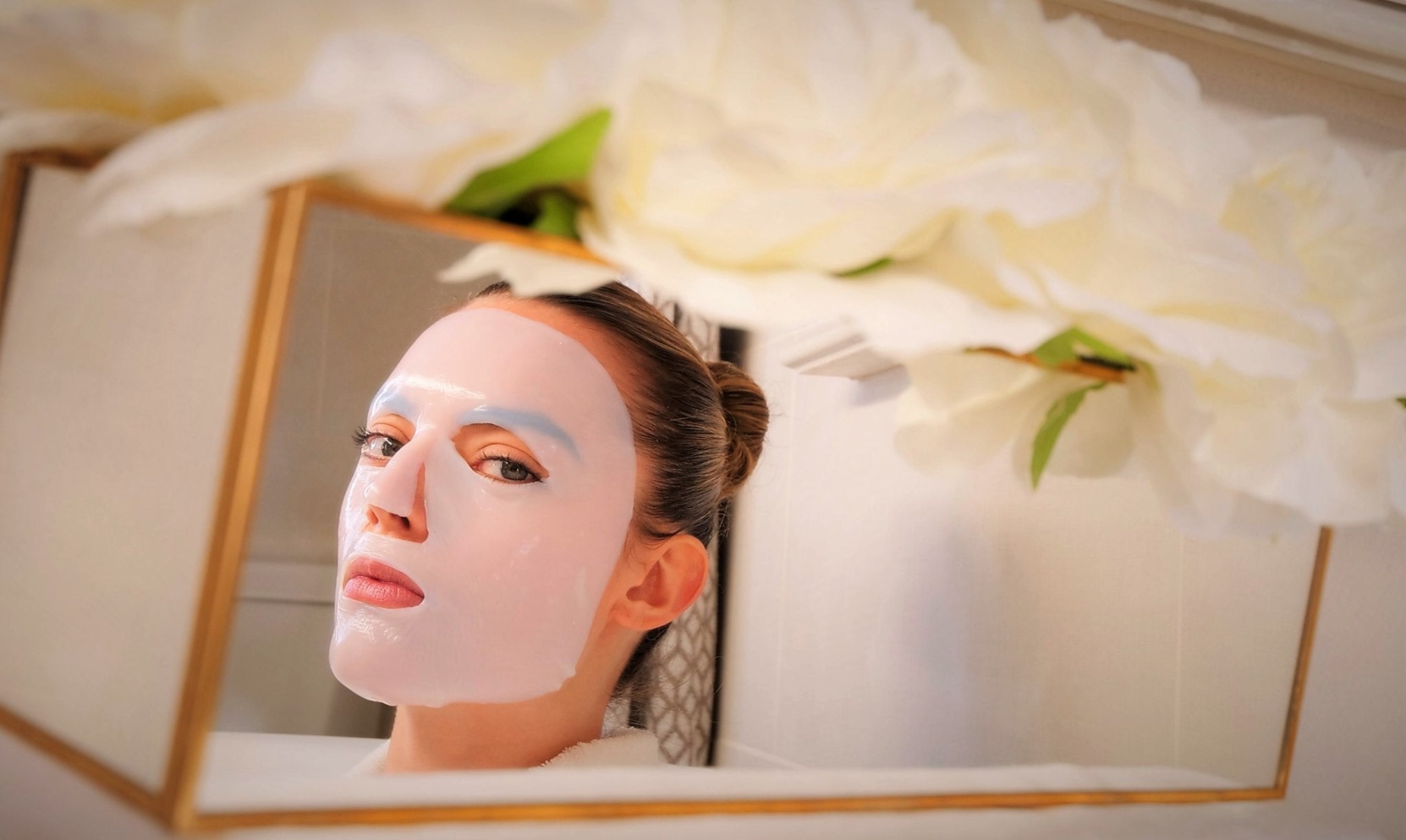 a woman wearing a skin care face mask looking into the mirror with flowers around her