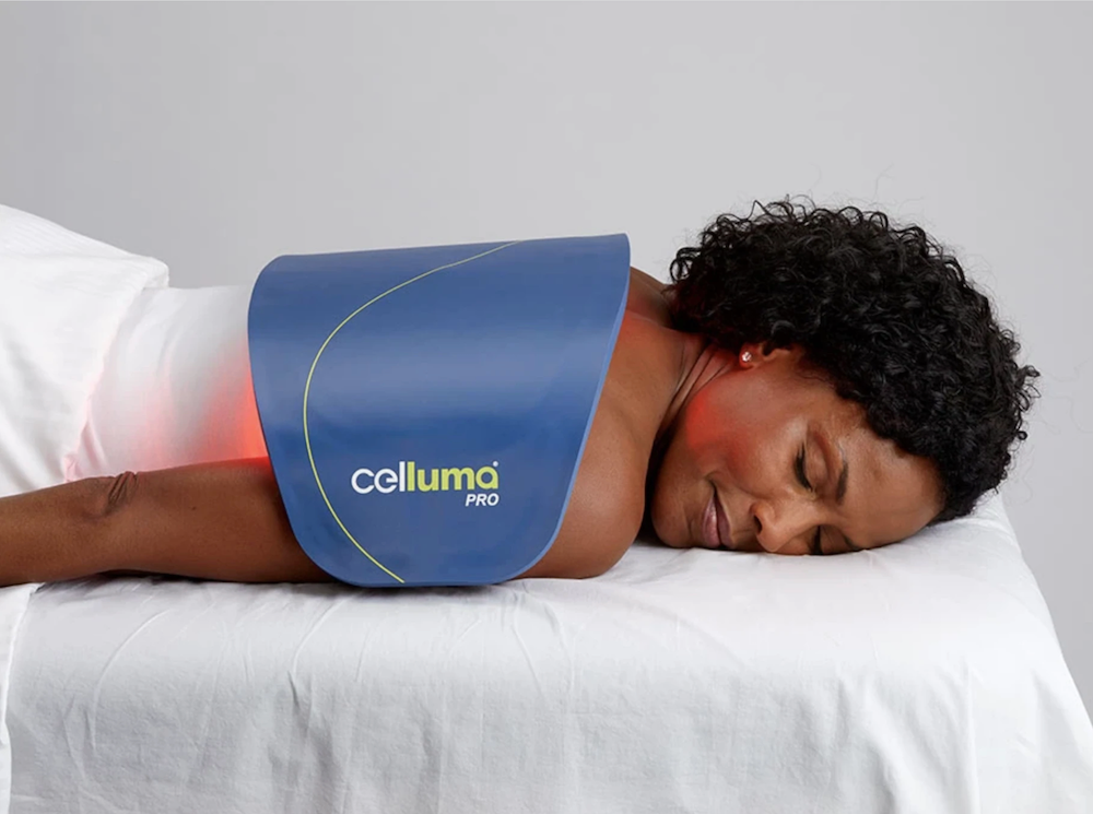 woman lying on a spa treatment table with the Celluma LED Light Therapy device across her back. The device is on and glowing red.
