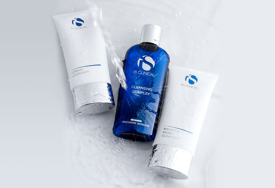 flat lay of three iS Clinical cleansers with water being poured onto them. the products include cream cleanser, cleansing complex, and warming honey cleanser