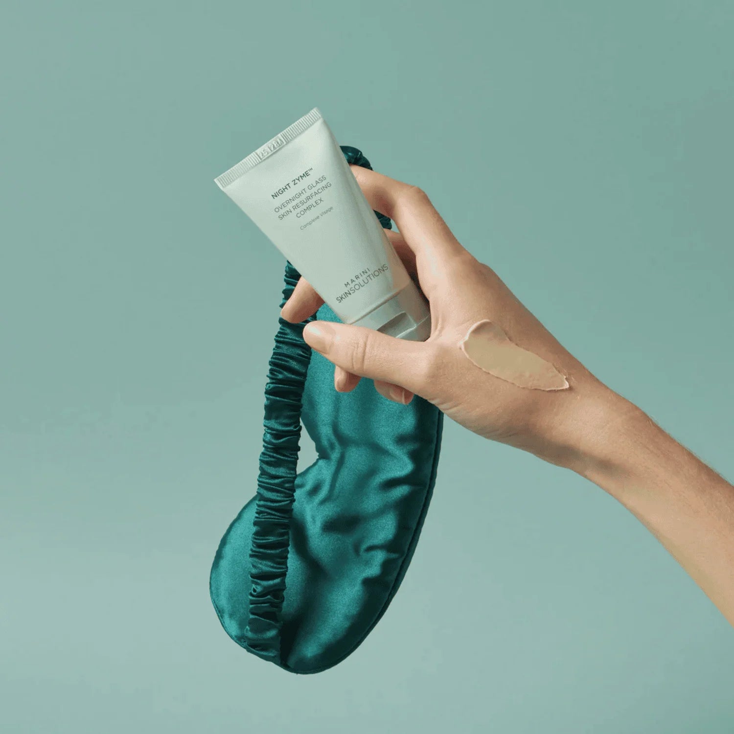 Hand holding a tube of Night Zyme with a green satin eye mask on a light green background